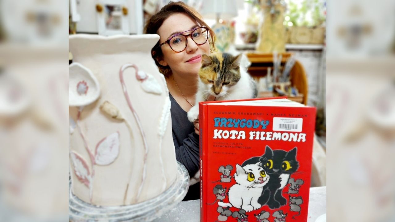 Na pierwszym planie książka "Przygody kota Filemona", a obok wazon gliniany. Na drugim planie kobieta w okularach trzyma w dłoniacah kota.