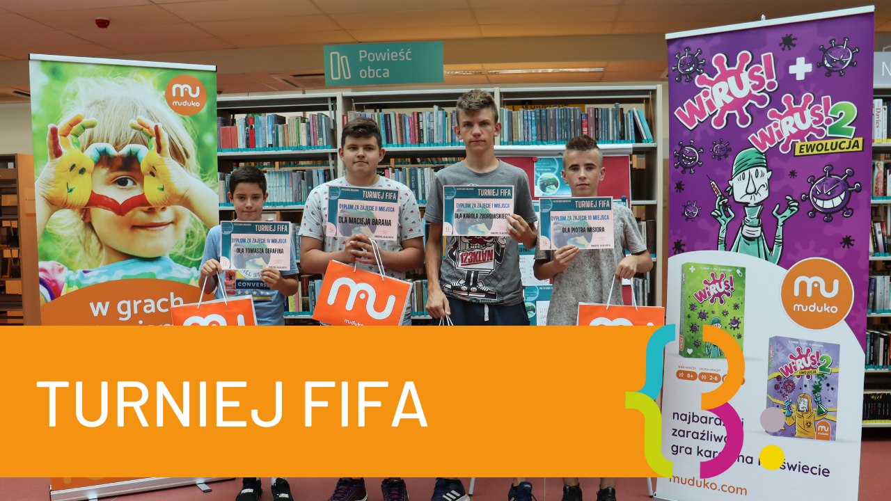Czworo chłopców stoi w bibliotece z dyplomami Turnieju Fifa i torebkami firmy Muduko. Obok nich, po obu bannery reklamujące firmę "Muduko" i grę "Wirus 2 Ewolucja". W tle regały z książkami. Napis Turniej FIFA