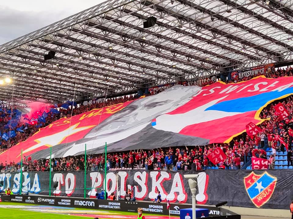 Na zdjęciu znajduje się sektor kibiców na stadionie. Na pierwszym planie znajduje się wielki, czarny banner z napisem "Wisła to Polska!", oraz godłem klubu Wisły. Na drugim planie tłum kibiców ubrany w większości na czerwono, trzyma ogromne, czerwone płótno z postacią mężczyzny w garniturze i napisami "KS Wisła" i "TS Wisła"