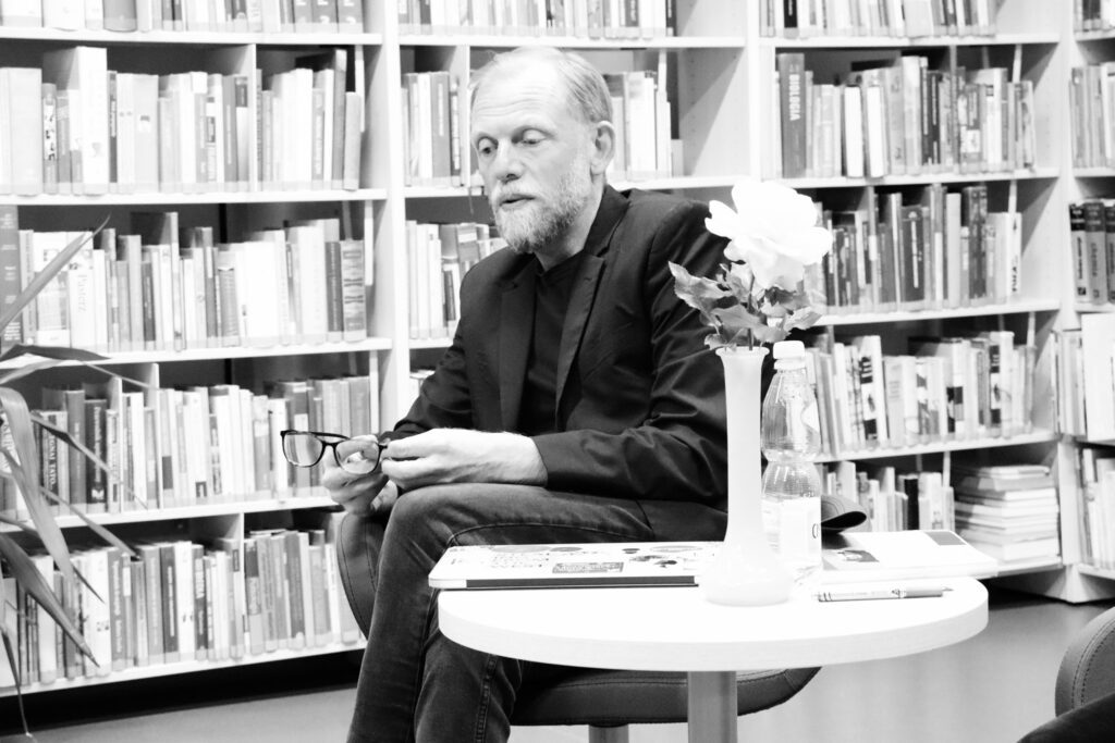 Autor Max Cegielski siedzący przy stoliku i opowiadający historię. Trzyma w rękach czarne okulary. W tle regały z książkami.