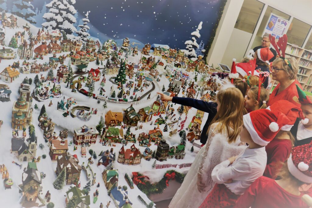 Grupa ludzi w tym dorośli i dzieci oglądają makietę świątecznego miasteczka w bibliotece. Jedno dziecko wskazuje palcem na makietę. Wszyscy przebrani są mikołajkowo, mają czapki czerwone albo rogi reniferów.
