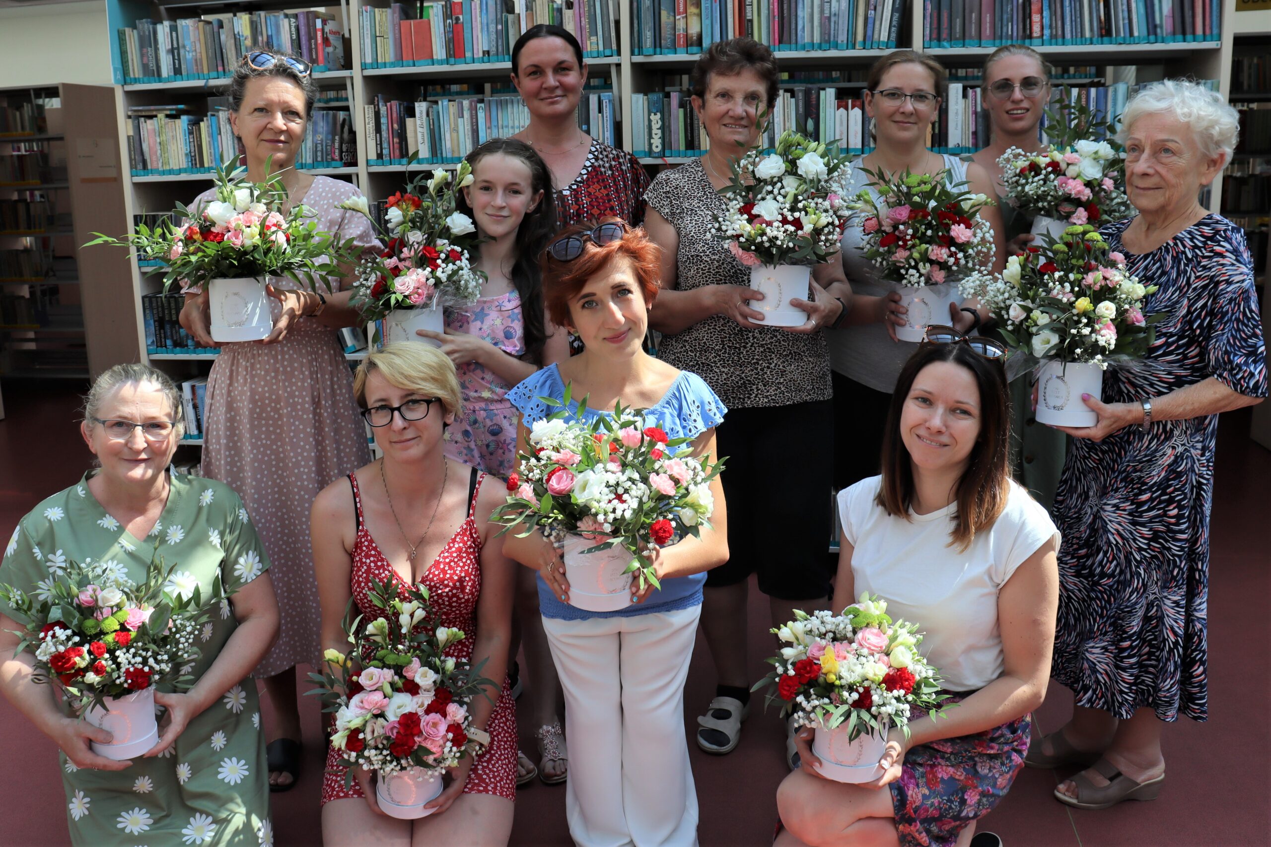 Jedenaście kobiet uśmiechniętych kobiet pozuje do zdjęcia w bibliotece. Na rękach trzymają wykonane przez siebie kompozycje florystyczne. 