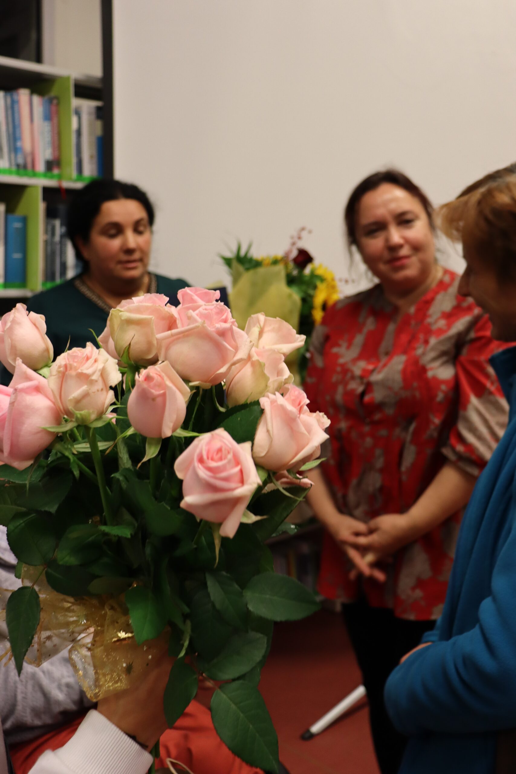 Na pierwszym planie wyciągnięta dłoń trzyma bukiet róż. Na drugim planie trzy kobiety rozmawiające ze sobą kobiety. Jedna z kobiet stojąca po prawej stronie trzyma w dłoniach bukiet kwiatów.