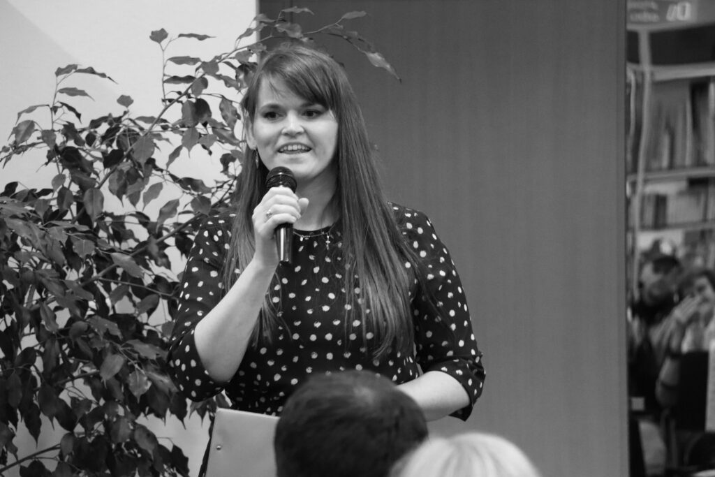 Młoda kobieta z uśmiechem spogląda na publiczność przed sobą i wygłasza do mikrofonu swoje przemówienie.