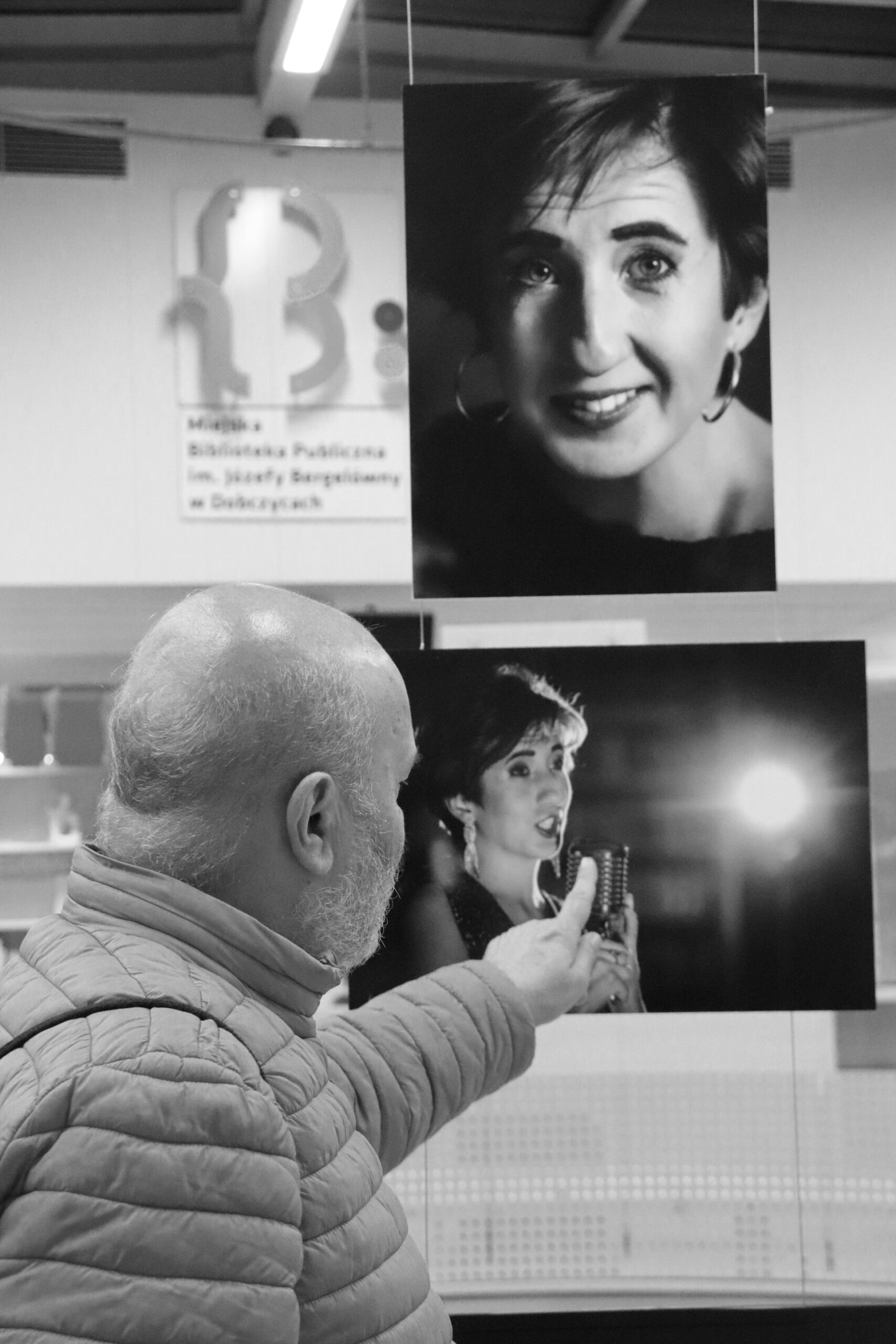 Stojący po lewej stronie mężczyzna, zwrócony tyłem do obiektywu wskazuje palcem na wiszące przed nim dwa portrety uczestniczki projektu. W tle na ścianie wisi logo Miejskiej Biblioteki Publicznej w Dobczycach.