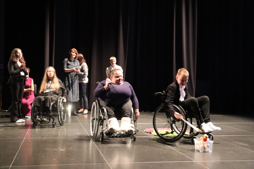 Na scenie pięć dorosłych osób i dwoje uczniów szkoły podstawowej bierze udział w prelekcji. Trzy osoby siedzą na wózku inwalidzkim