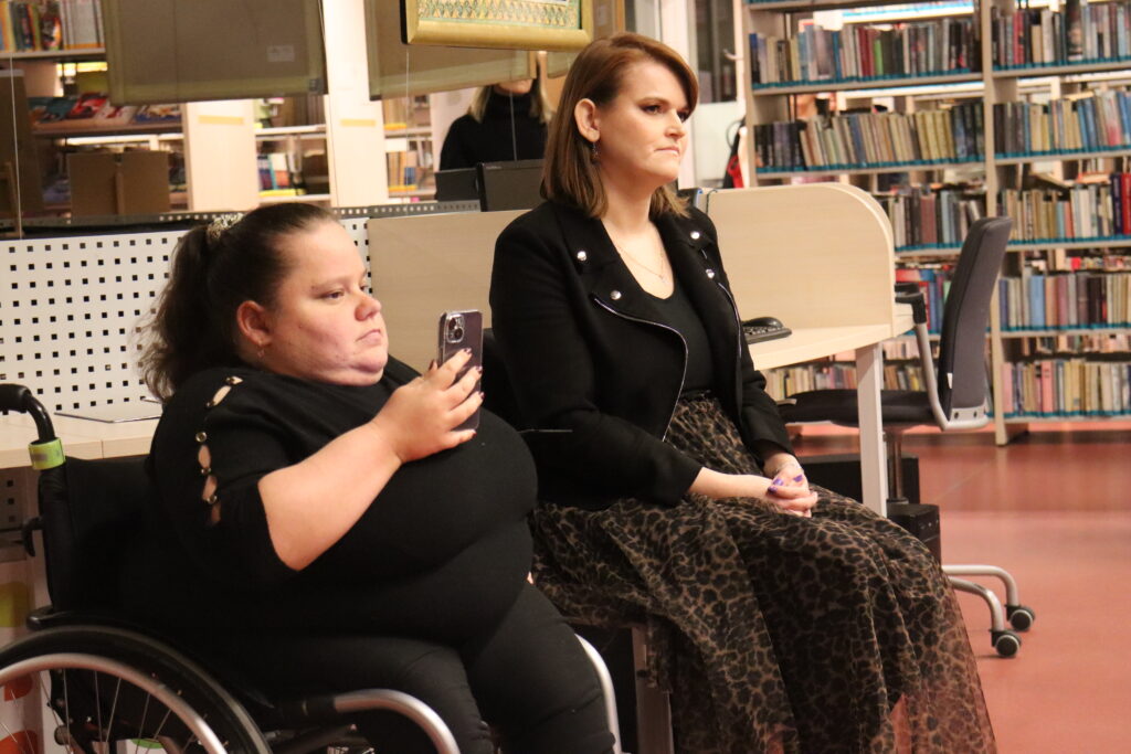Dwie kobiety słuchają i oglądają prelekcję. Jedna z nich porusza się na wózku i kameruje wystąpienie telefonem komórkowym