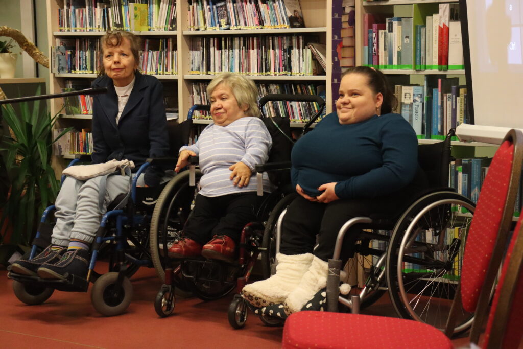 Trzy kobiety na wózku inwalidzkim uśmiechnięte siedzą, słuchając przemówienia.