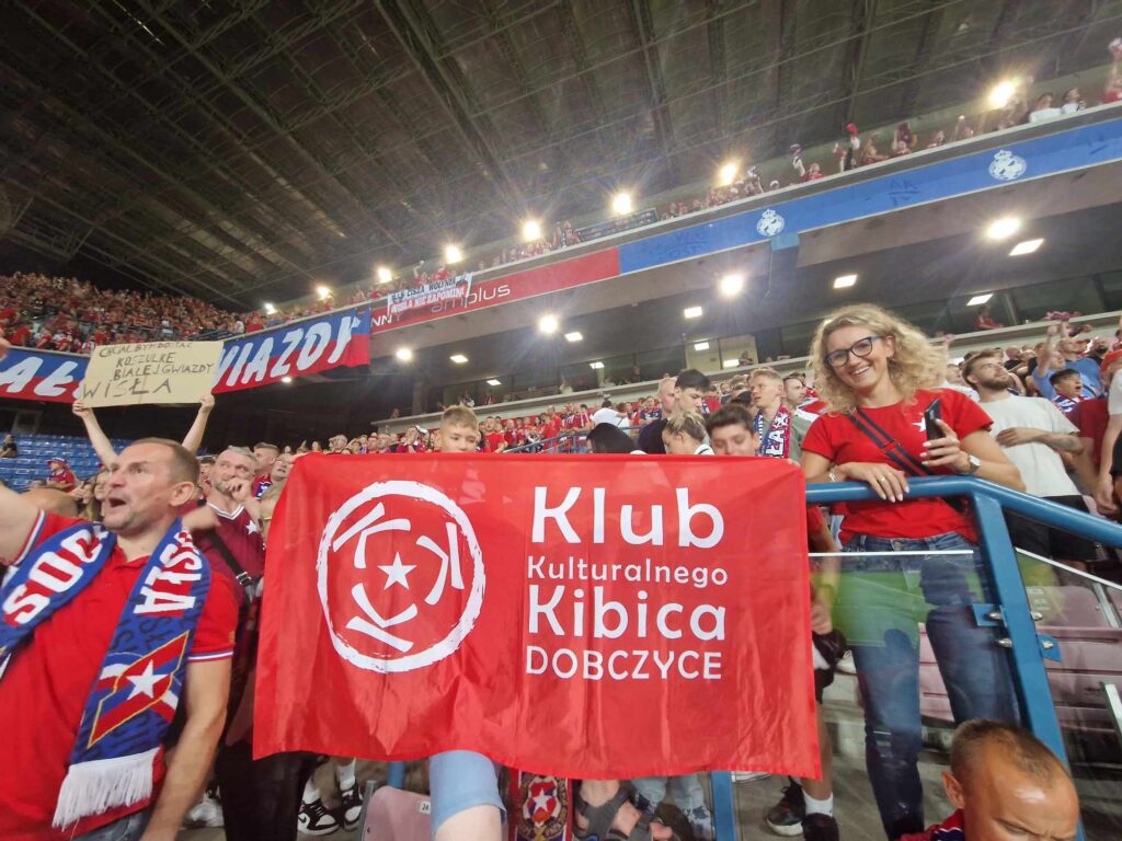 Grupa kibiców na trybunach pozuje z zawieszoną flagą Klubu Kulturalnego Kibica w kolorze czerwonym