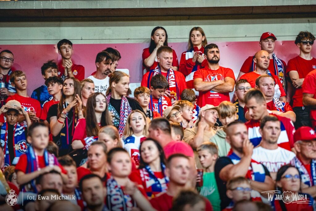 Grupa kibiców na trybunach piłkarskich pochłonięta jest kibicowaniem i oglądaniem meczu. Większość z nich ma na sobie czerwone koszulki i szaliki z barwami klubu Wisła Kraków,