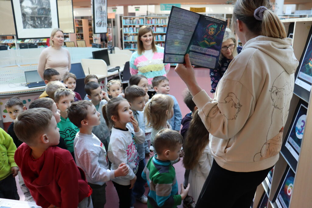 Grupa przedszkolaków w bibliotece słucha bibliotekarki, pokazującej im kolorową kartę na naklejki.