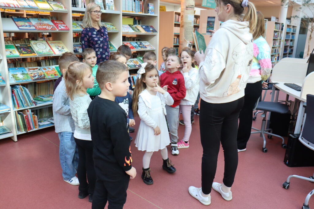 Grupa przedszkolaków w bibliotece słucha bibliotekarki.