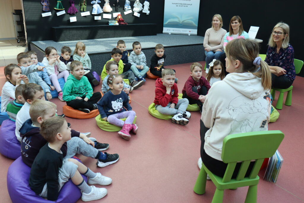 Przedszkolaki siedzą na kolorowych pufach i słuchają książki, czytanej im przez bibliotekarkę.