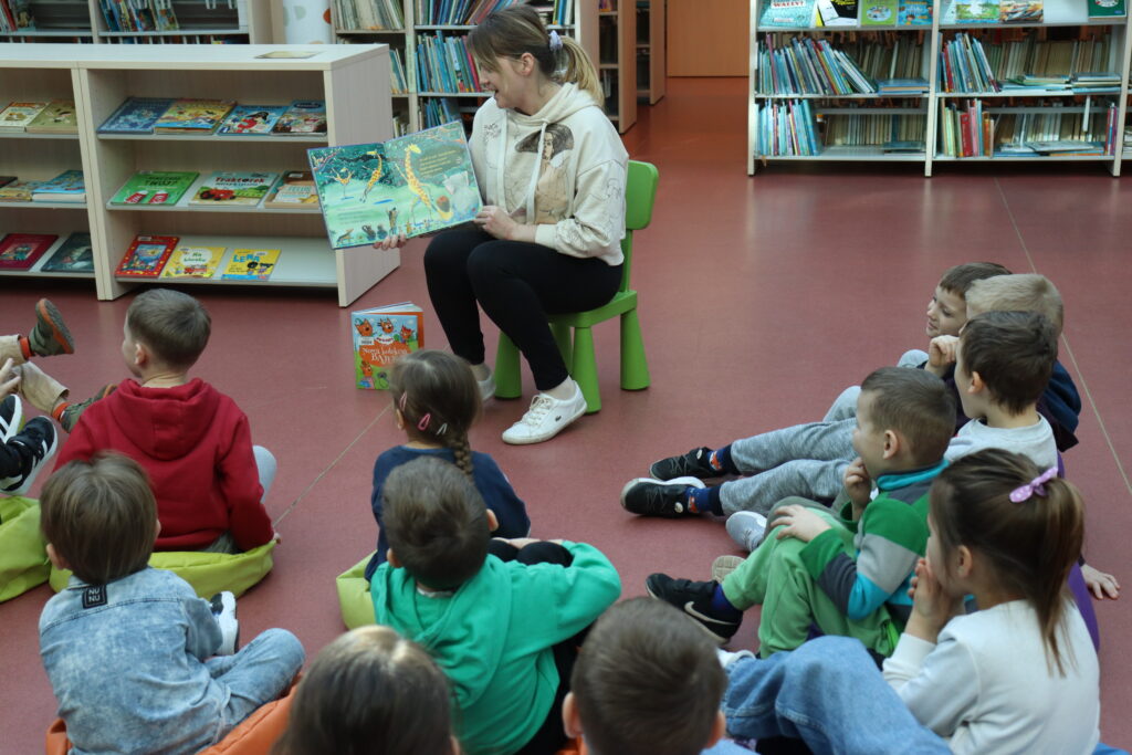 Przedszkolaki siedzą na kolorowych pufach i słuchają książki, czytanej im przez bibliotekarkę.