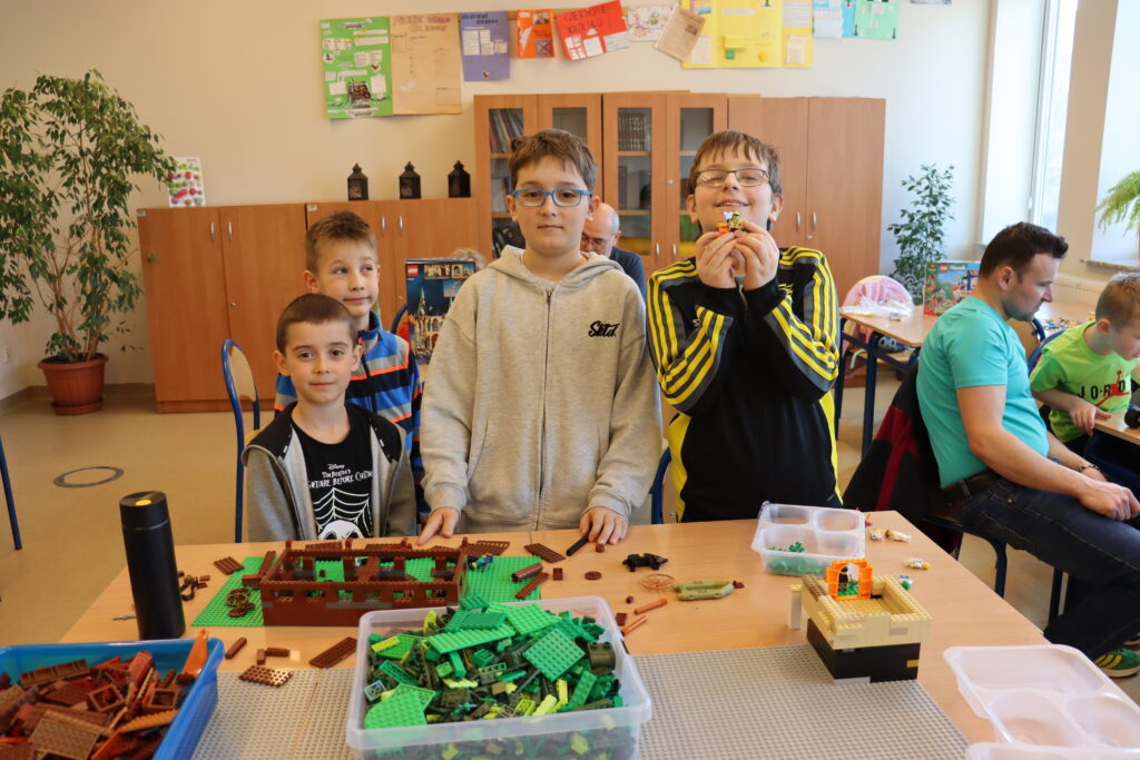Czterech chłopców w sportowych ubraniach stoi przy stole, na którym rozłożone są zielone i brązowe klocki, z których budują stajnię.