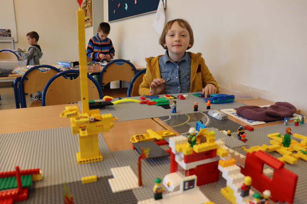 Chłopiec w niebieskiej koszuli i żółtej bluzie siedzi przy stole, przed nim zbudowana przez niego żółta, wysoka wieżą i kolorowy domek.