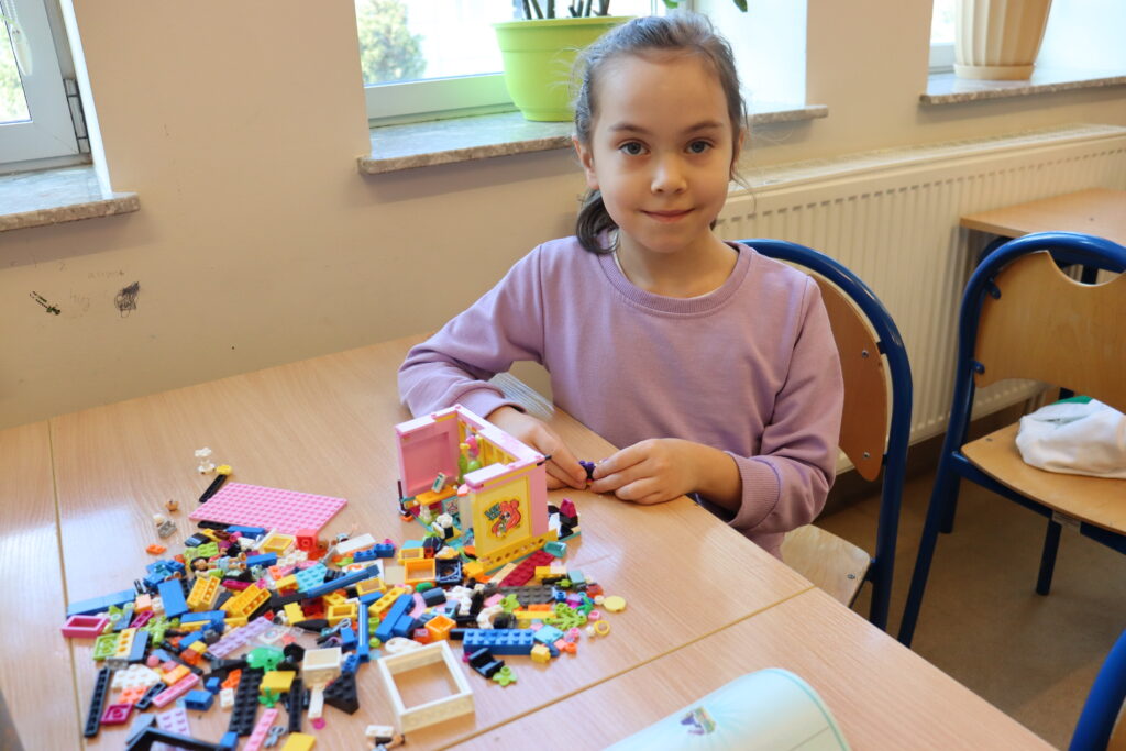 Dziewczynka w różowej bluzie buduje domek z klocków.