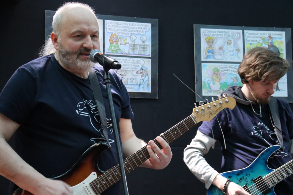 Dwóch członków zespołu z gitarami elektrycznymi, po lewej starszy mężczyzna z siwą brodą i długimi włosami, po prawej mężczyzna w granatowej bluzie.