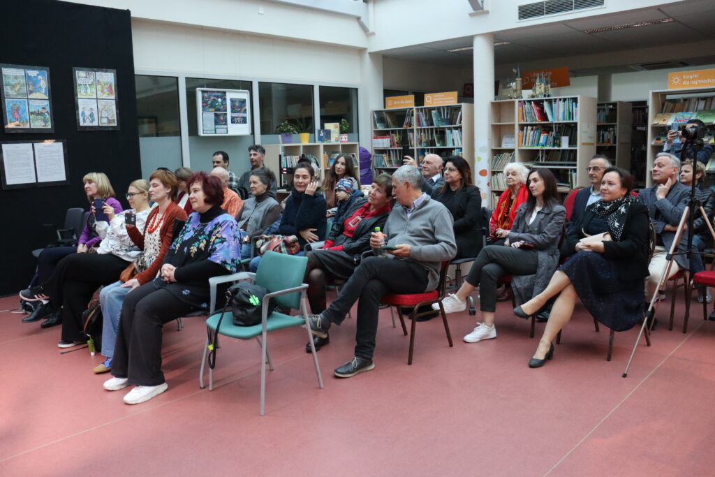 Publiczność zgromadzona podczas koncertu siedzi na krzesłach w przestrzeni biblioteki.