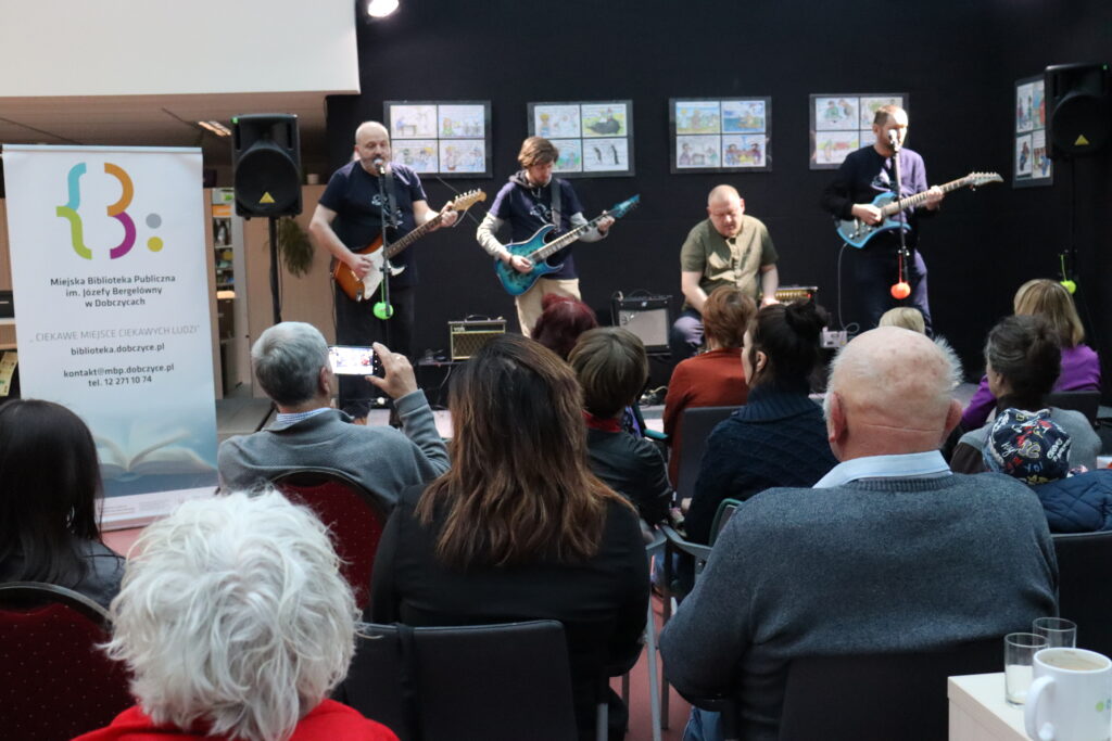 Publiczność zgromadzona podczas koncertu siedzi na krzesłach w przestrzeni biblioteki; na scenie czterech muzyków z intrumentami.