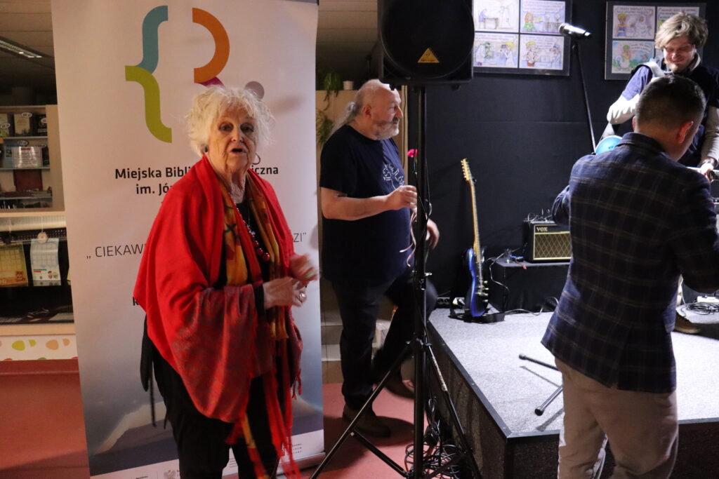 Aktorka Magdalena Sokołowska w czerwonej narzutce pozuje na tle baneru biblioteki.