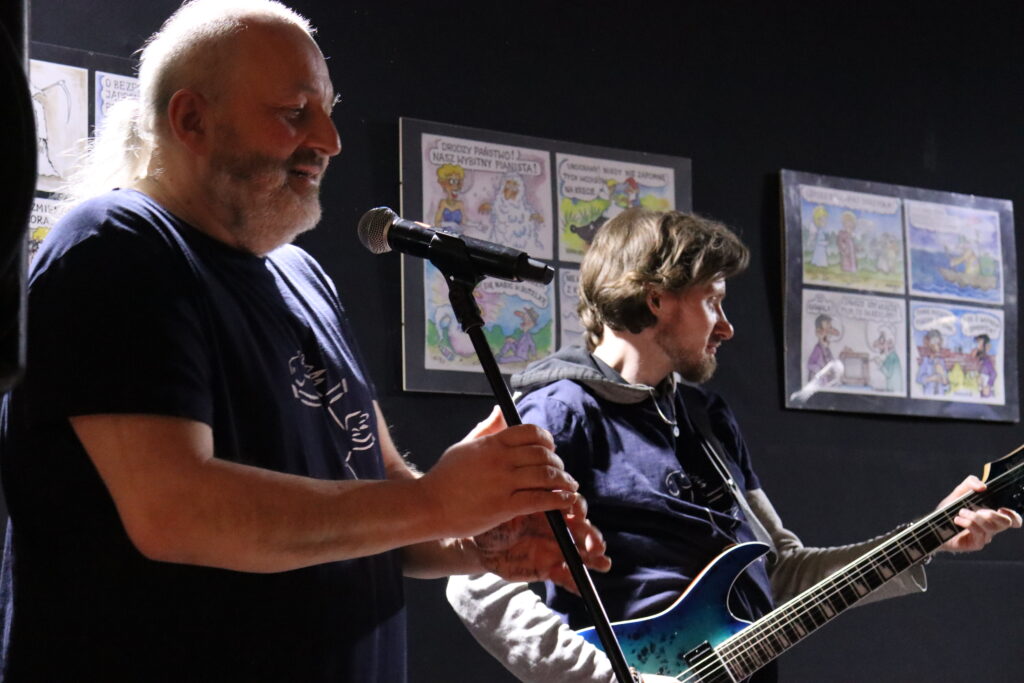 Dwóch członków zespołu: po lewej starszy mężczyzna z siwą brodą i długimi włosami trzyma mikrofon, po prawej mężczyzna w granatowej bluzie z gitarą elektryczną.