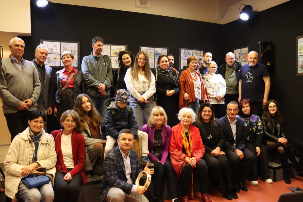 Członkowie zespołu oraz goście wydarzenia pozują do grupowego zdjęcia na scenie w bibliotece.