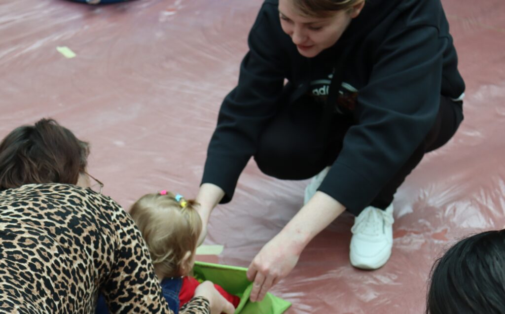 Zbliżenie na kobietę w czarnej bluzie i drugą osobę w panterkę, które pomagają małemu dziecku bawić się przy zielonym materiale na podłodze wyłożonej folią.