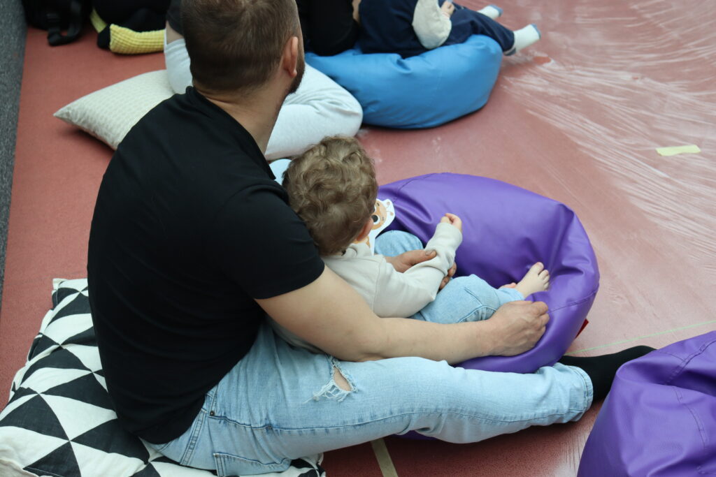 Mężczyzna w czarnym t-shircie i jasnych dżinsach siedzi na podłodze, trzymając na kolanach małe dziecko o kręconych włosach; obok nich leżą kolorowe pufy i poduszki.