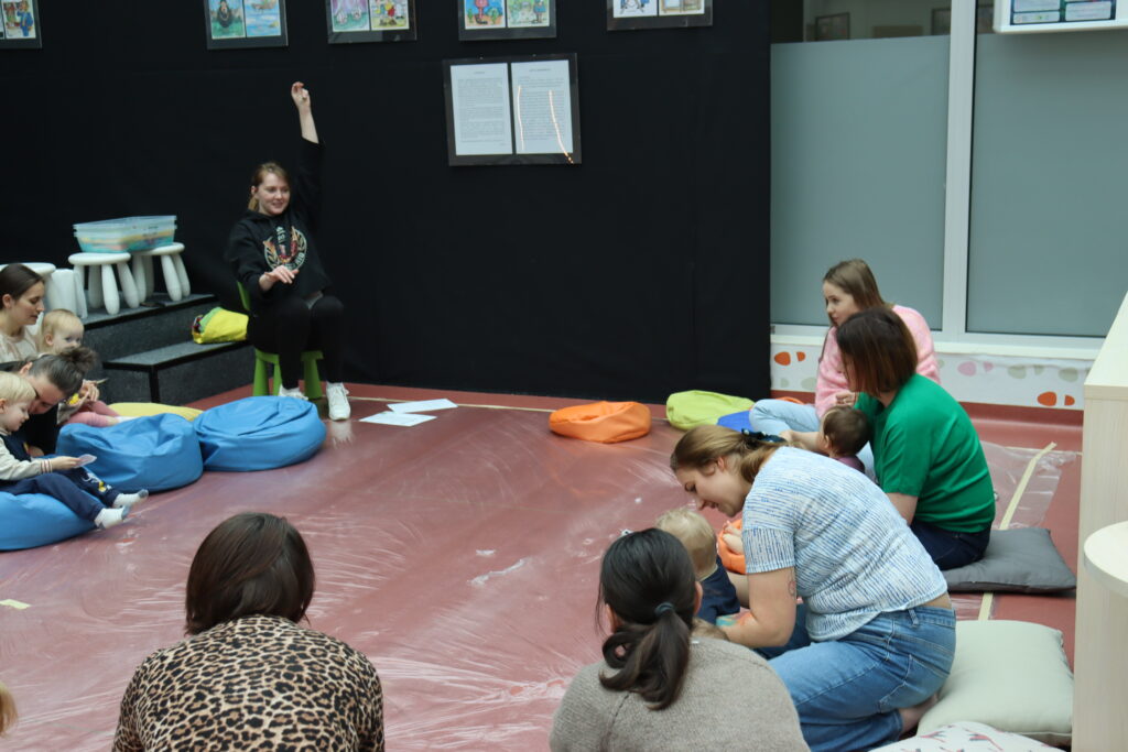 Grupa rodziców z małymi dziećmi siedzi w kręgu na podłodze w nowoczesnej sali; prowadząca w czarnej bluzie unosi rękę, inicjując zabawę.