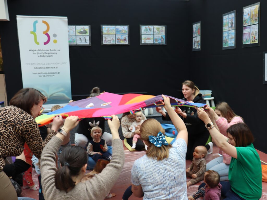 Rodzice i dzieci wspólnie trzymają brzegi dużej, wielokolorowej chusty animacyjnej, unosząc ją nad siedzącymi na środku maluchami.