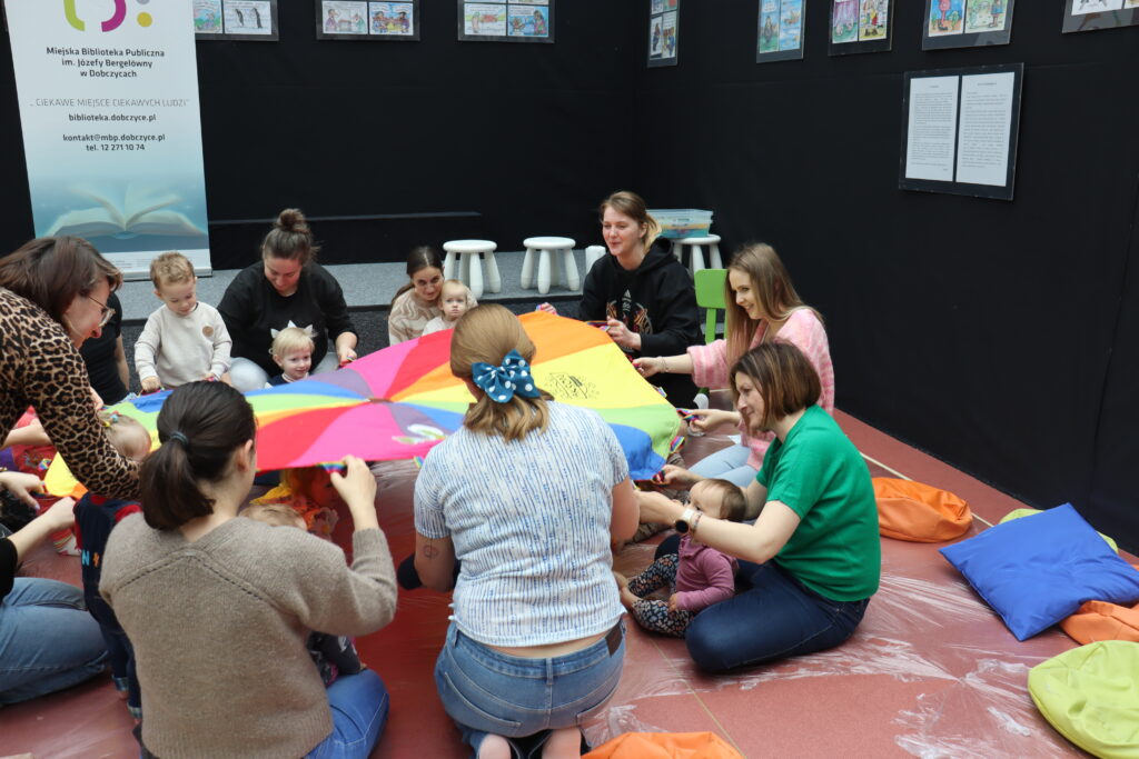 Grupa osób siedzi w kręgu i trzyma napiętą kolorową chustę; w tle widać baner informacyjny Miejskiej Biblioteki Publicznej w Dobczycach.