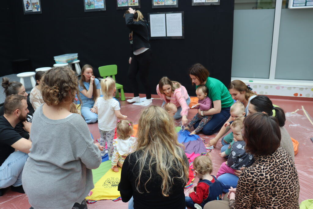 Dzieci i dorośli zgromadzeni wokół kolorowej chusty rozłożonej na podłodze; maluchy raczkują i chodzą po materiale pod opieką rodziców.