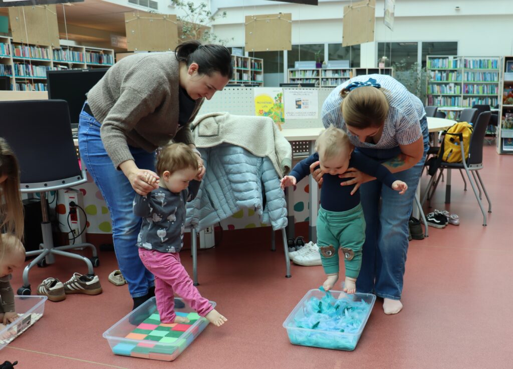 Dwie kobiety pomagają małym dzieciom stawiać pierwsze kroki w plastikowych pojemnikach wypełnionych różnymi fakturami: kolorowymi gąbkami oraz niebieskimi piórami.