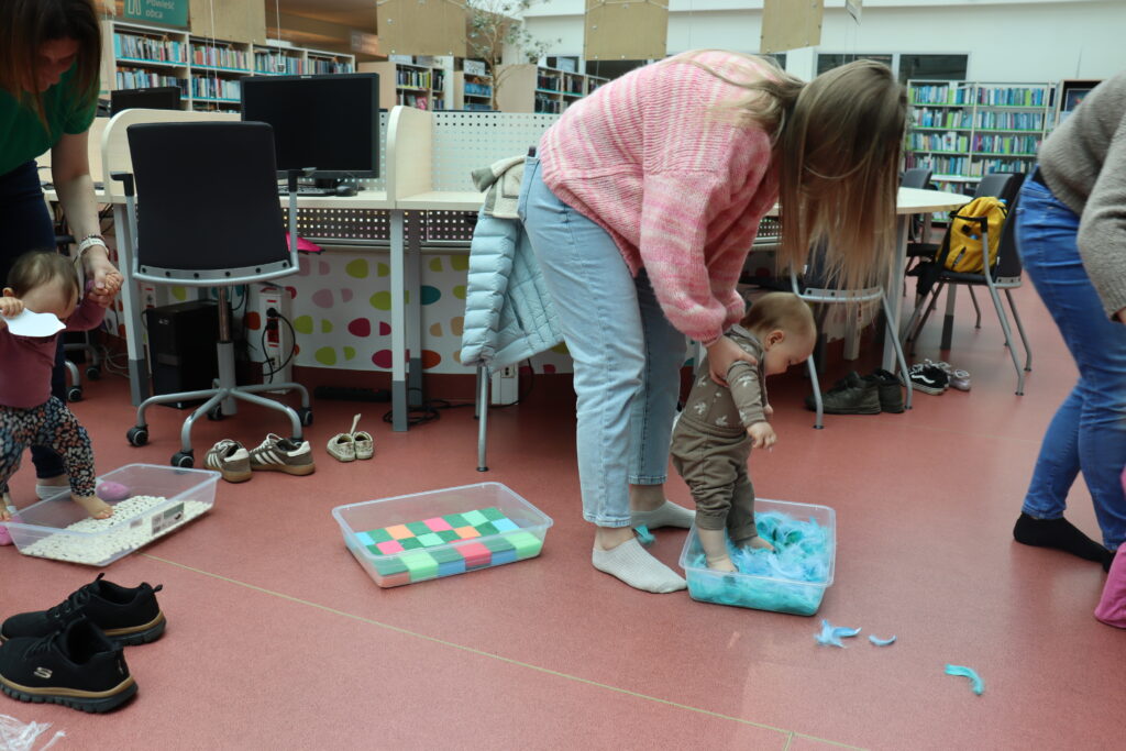 Małe dziecko podtrzymywane przez opiekuna stoi boso w przezroczystym pudełku wypełnionym niebieskimi piórami; obok znajduje się drugie pudełko z kolorowymi gąbkami.