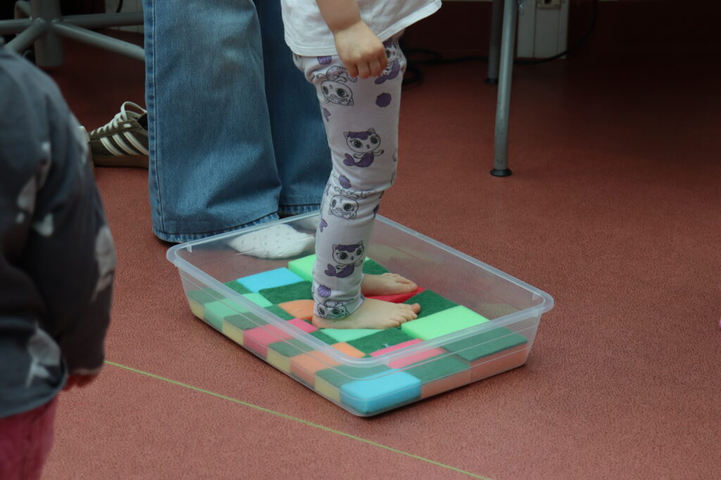 Zbliżenie na bose stopy dziecka stojącego w plastikowym pojemniku wypełnionym wielokolorowymi, miękkimi gąbkami do zmywania.