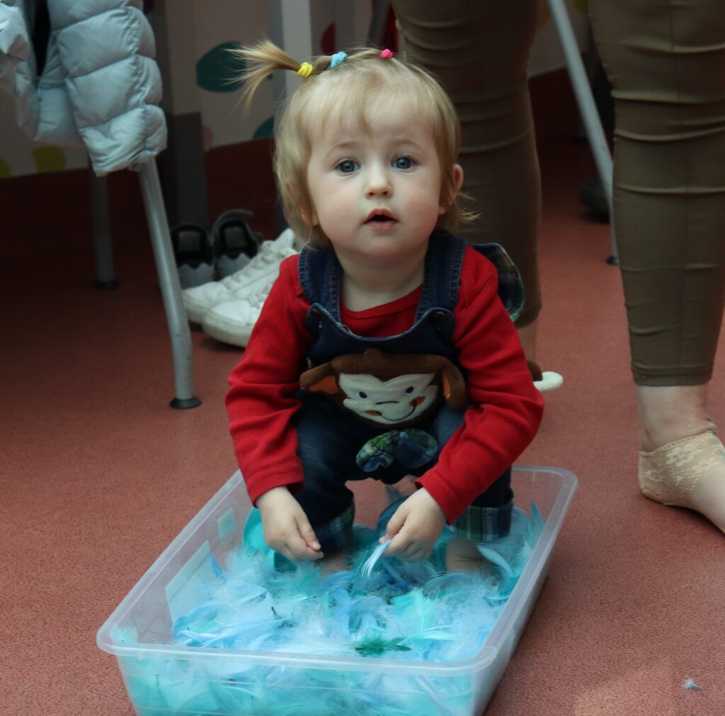 Mała dziewczynka w czerwonej bluzce i ogrodniczkach z małpką kuca w przezroczystym pojemniku pełnym jasnoniebieskich piór, patrząc w stronę aparatu.