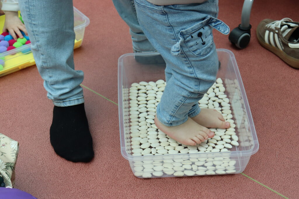 Zbliżenie na bose stopy dziecka stojącego w pojemniku wypełnionym białymi fasolkami, co stymuluje zmysł dotyku.