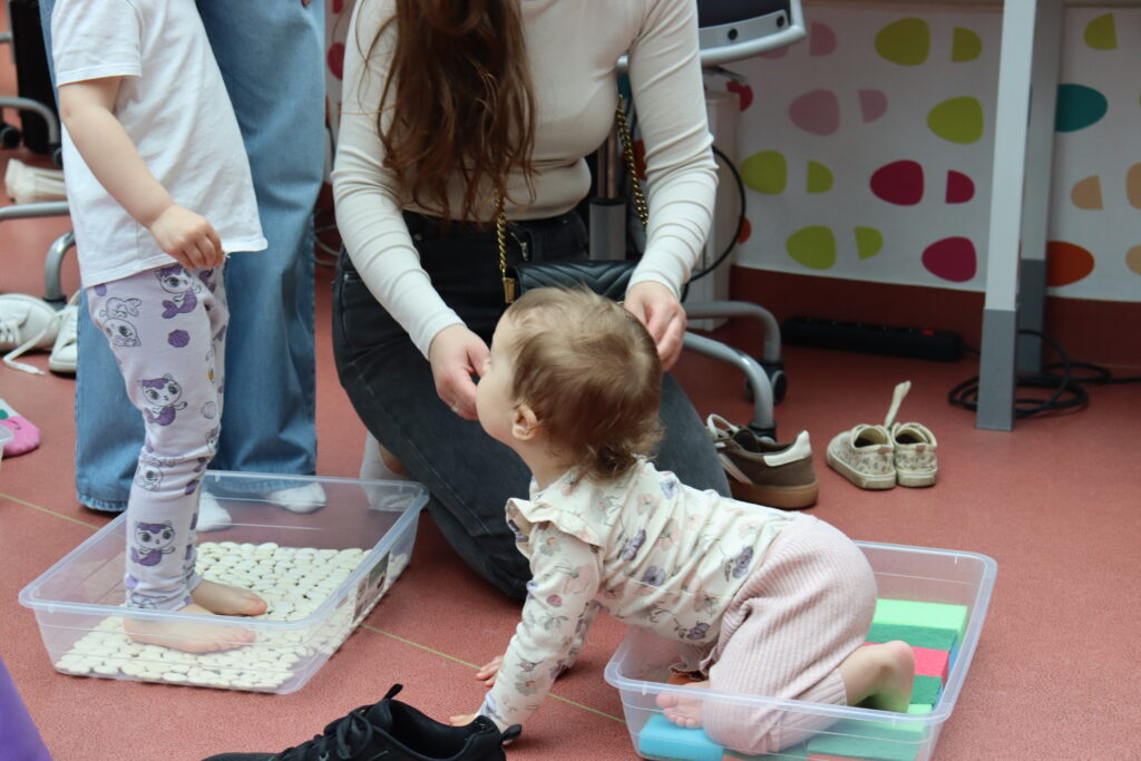 Małe dziecko bawi się niebieskimi piórami, siedząc w przezroczystym pojemniku na podłodze biblioteki.