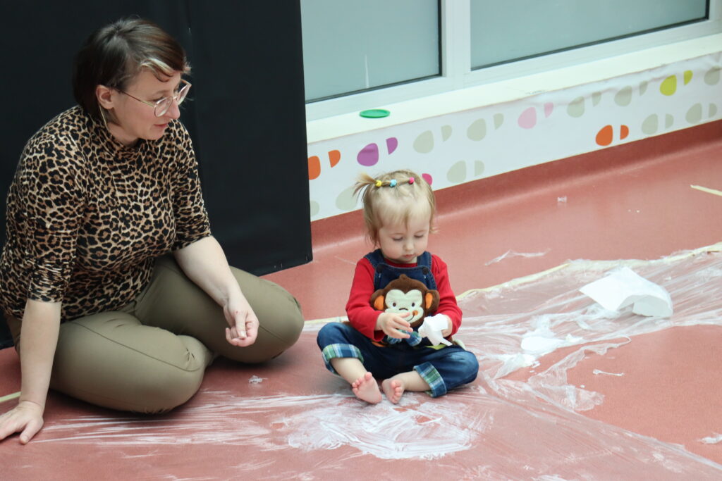 Kobieta w bluzce w panterkę obserwuje zabawy śmietanką małej dziewczynki siedzącej na folii malarskiej.