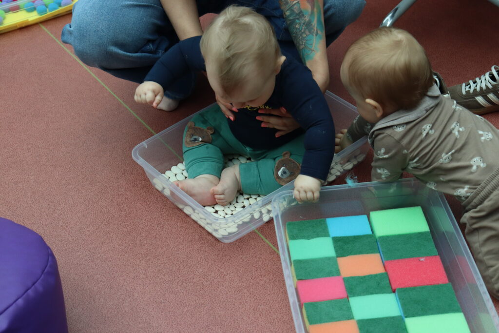 Maluch siedzi na podłodze biblioteki i bawi się białymi fasolkami rozsypanymi w przezroczystym pojemniku. obok drugi pojemnik z gąbkami.