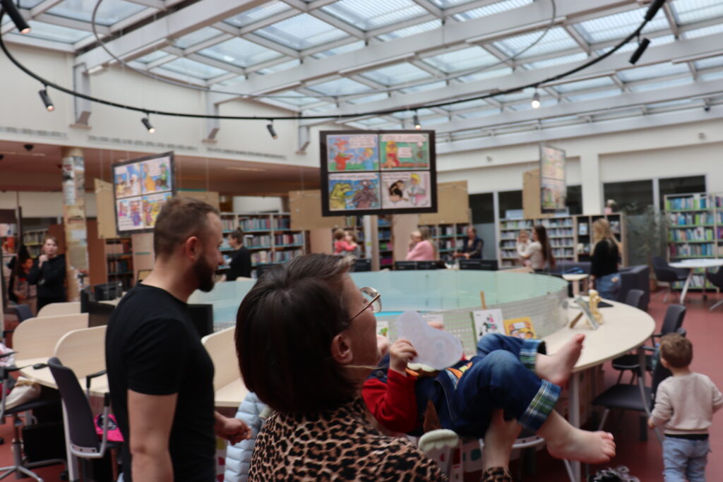 Kobieta podrzuca dzieckiem; w tle widoczne są regały z książkami i stanowiska komputerowe.