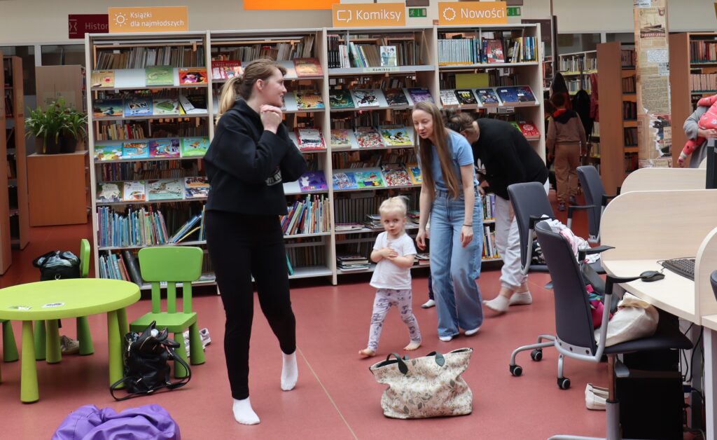 Grupa rodziców i dzieci na zakończenie zajęć w bibliotece; niektórzy sprzątają rekwizyty, inni rozmawiają w swobodnej atmosferze.