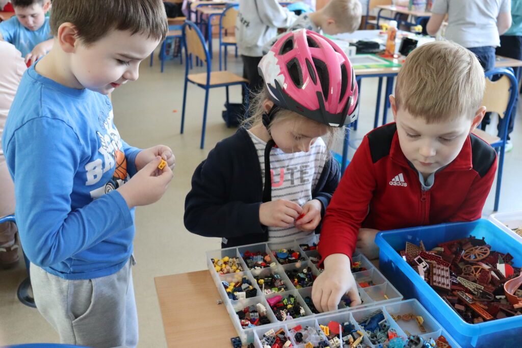 Dwóch chłopców i dziewczynka w różowym kasku szukają klocków w pojemnikach.
