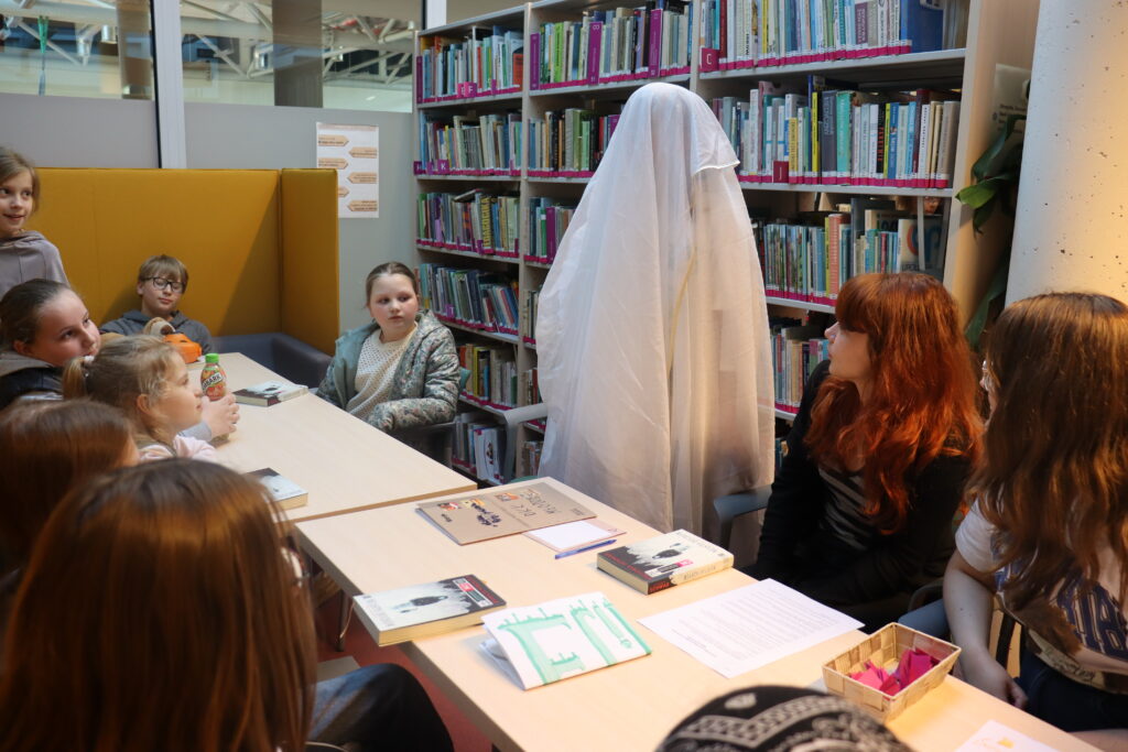 Dziewczynka w przebraniu ducha, przykryta tiulową firanką, stoi za stołem.