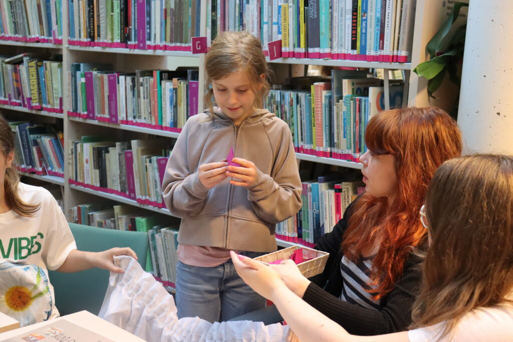 Dziewczynka w szarej bluzie otwiera małą fioletową karteczkę.