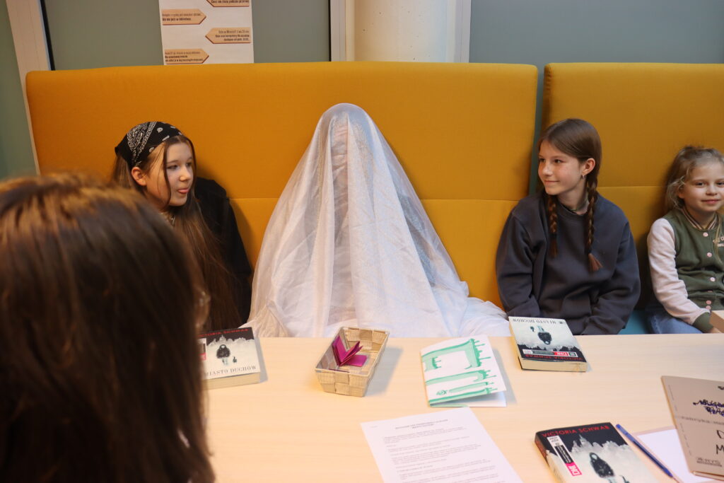 Dziewczynka w przebraniu ducha, przykryta tiulową firanką, siedzi za stołem. Po jej dwóch stronach siedzą dwie dziewczynki i się uśmiechają.