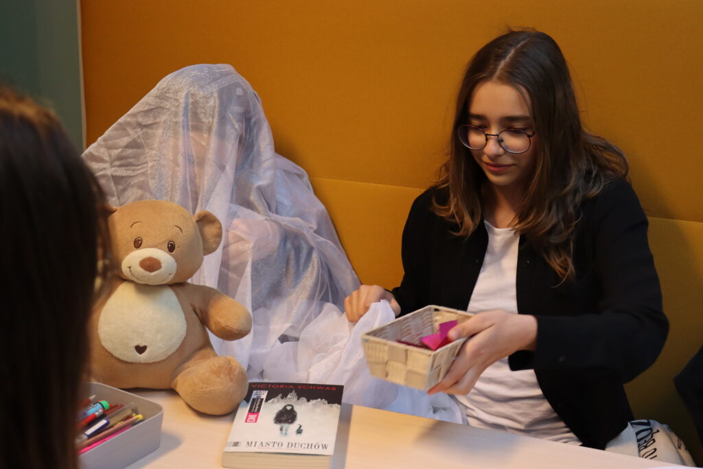 Dziewczynka z okularach sięga po pudełko z karteczkami; obok dziewczynka przykryta tiulową firanką trzyma pluszowego misia.