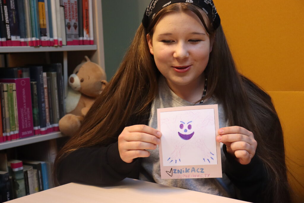 Dziewczynka z długimi ciemnymi włosami prezentuje swój rysunek: uśmiechnięty Duch Znikacz.