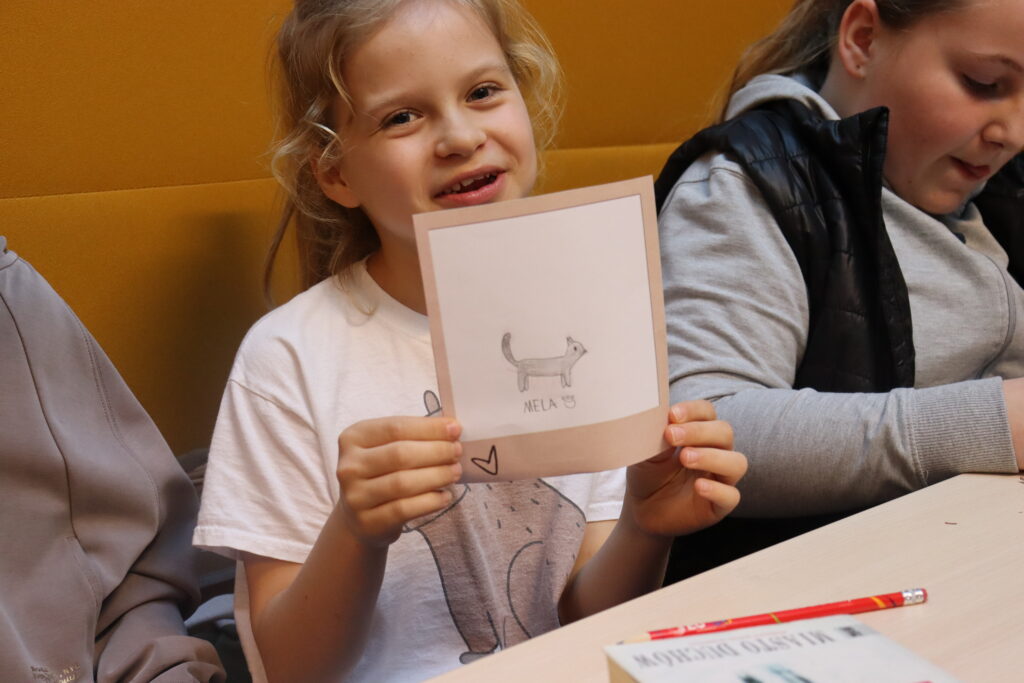 Dziewczynka z blond włosami prezentuje rysunek kotka z podpisem: Mela.